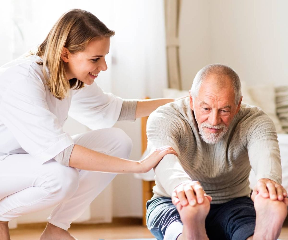 Health visitor and a senior man during home visit. A nurse or a physiotherapist helping a senior man exercise.