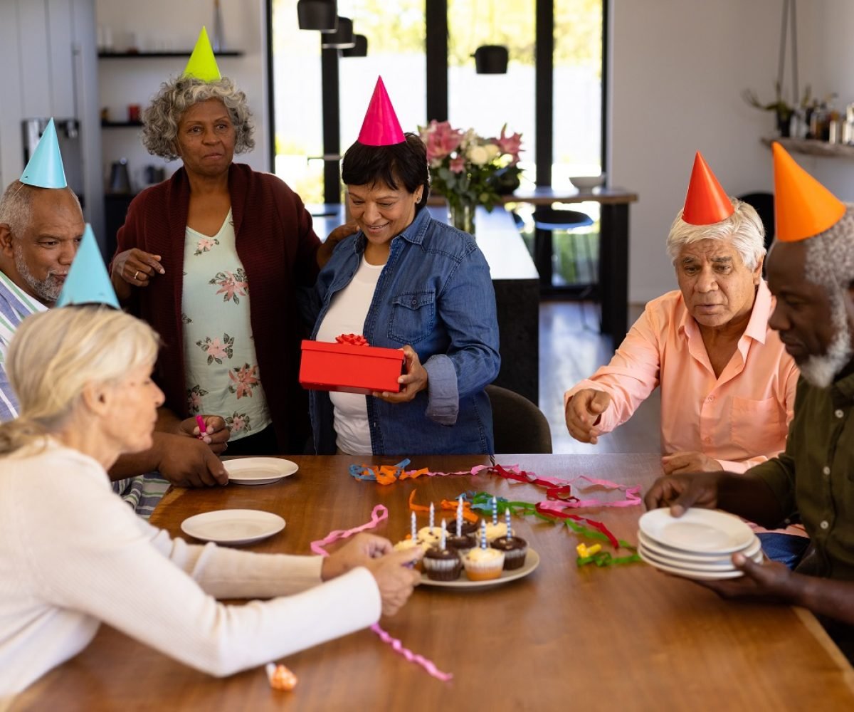 Multiracial senior friends wearing party hats doing preparation while woman holding birthday gift. Cupcake, plate, enjoyment, unaltered, togetherness, support, assisted living and retirement home.